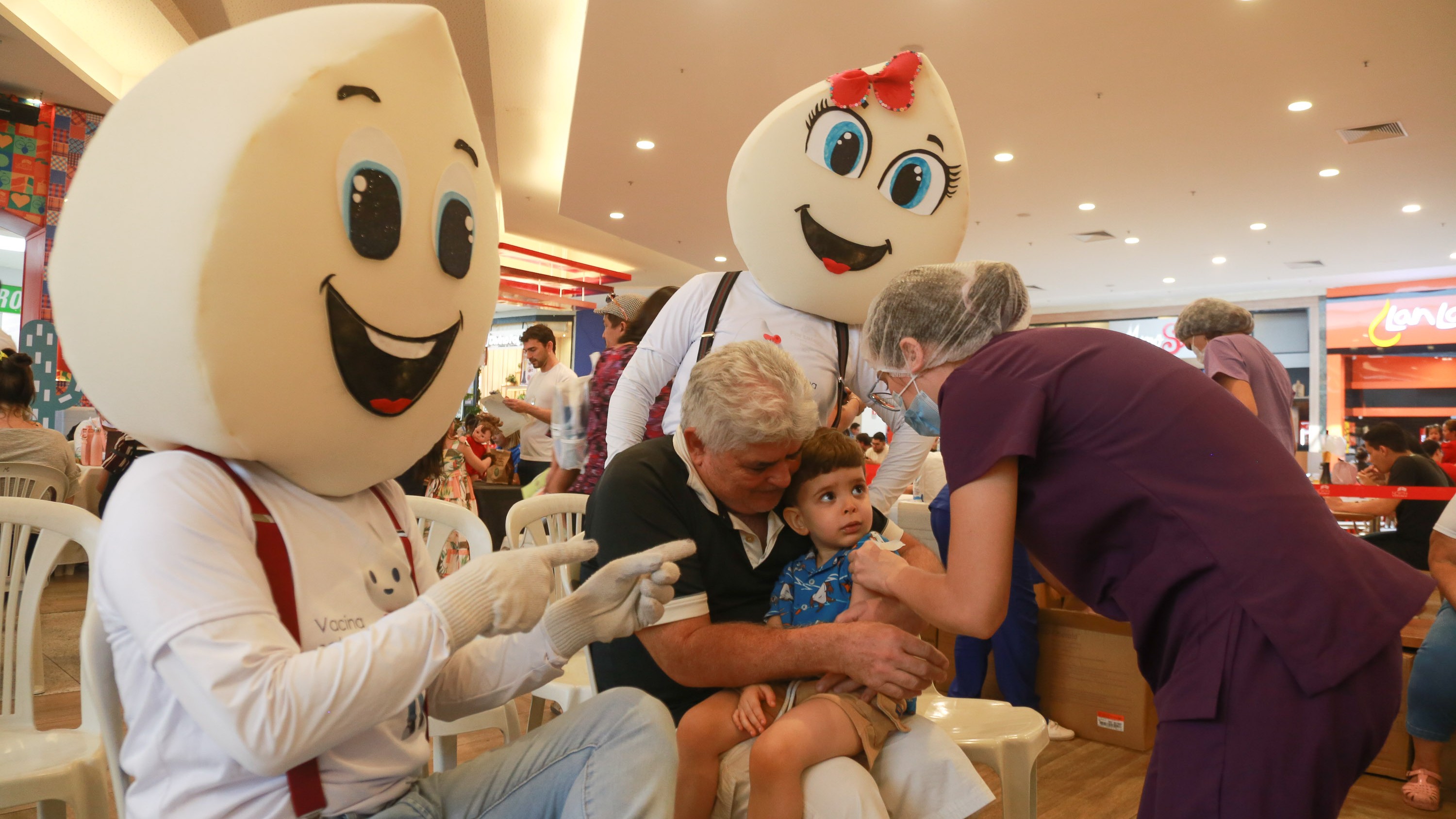 personagens vestidos de gota de vacina acenam para a câmera enquanto uma criança é vacinada no colo de um senhor